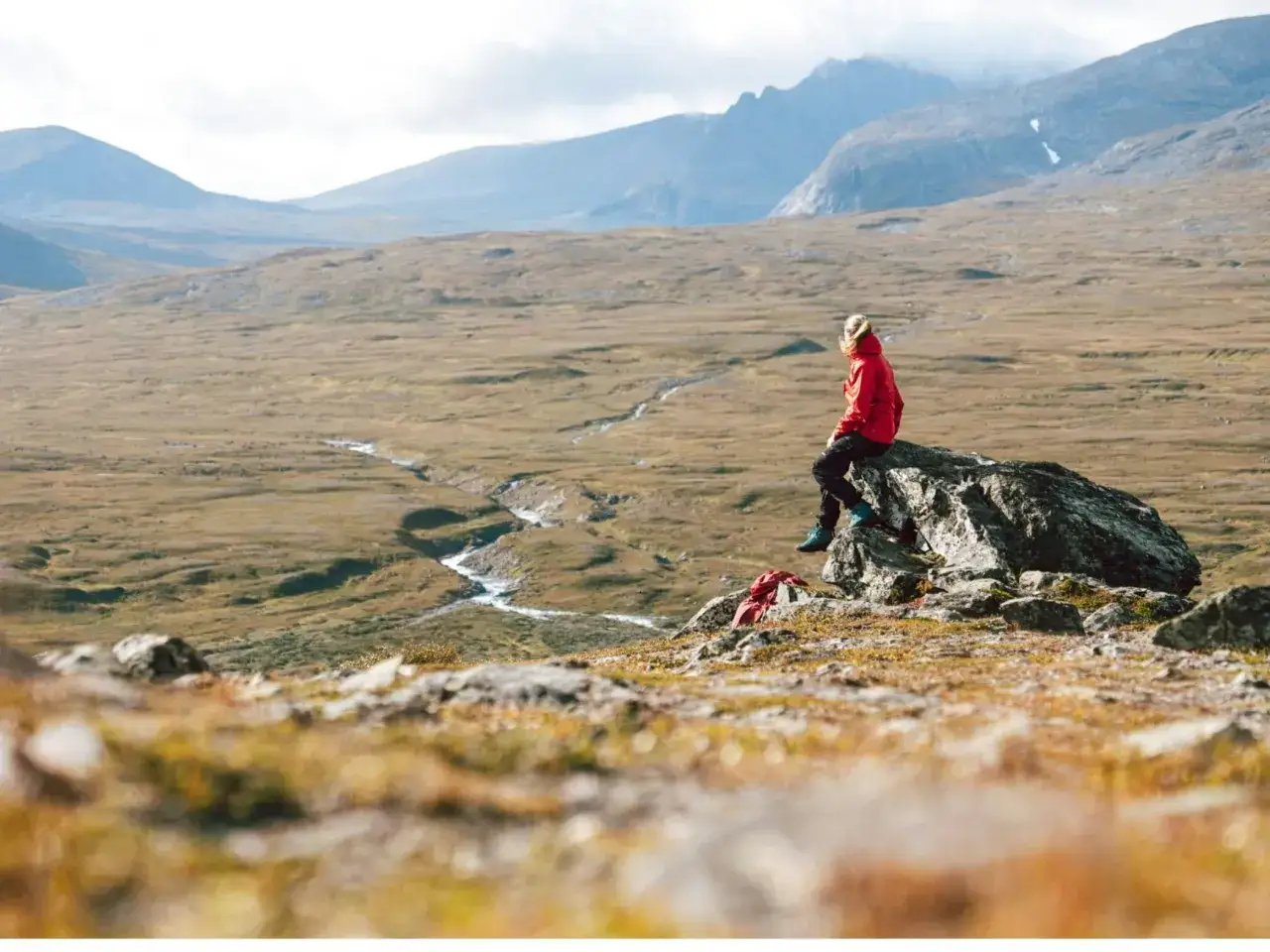 En person i r&ouml;d jacka vilar p&aring; en sten under en vandring i Storulv&aring;n. Vackert landskap med berg och en slingrande &auml;lv.