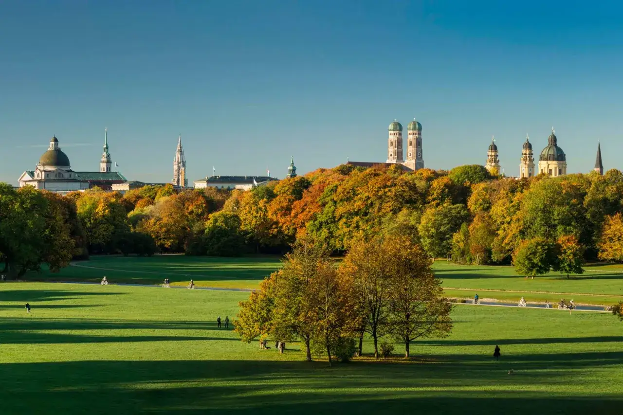 Herbstliche Farben im Englischen Garten, Schwabing, München. Menschen genießen die Sonne auf grünen Wiesen, während im Hintergrund die Skyline der Stadt mit ihren Türmen und Kuppeln aufragt.