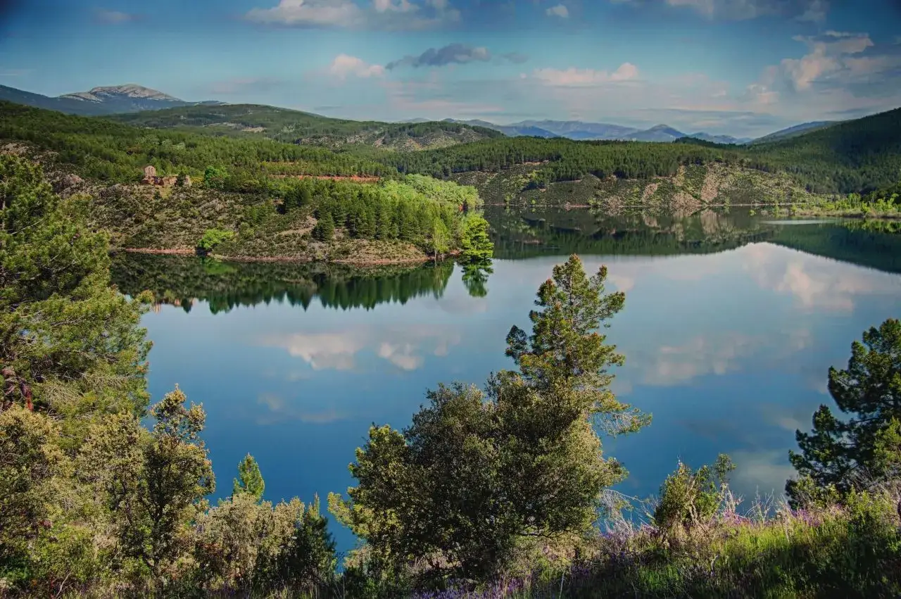 El embalse del Vado, rodeado de pinos y monta&ntilde;as, refleja un cielo azul con nubes blancas.