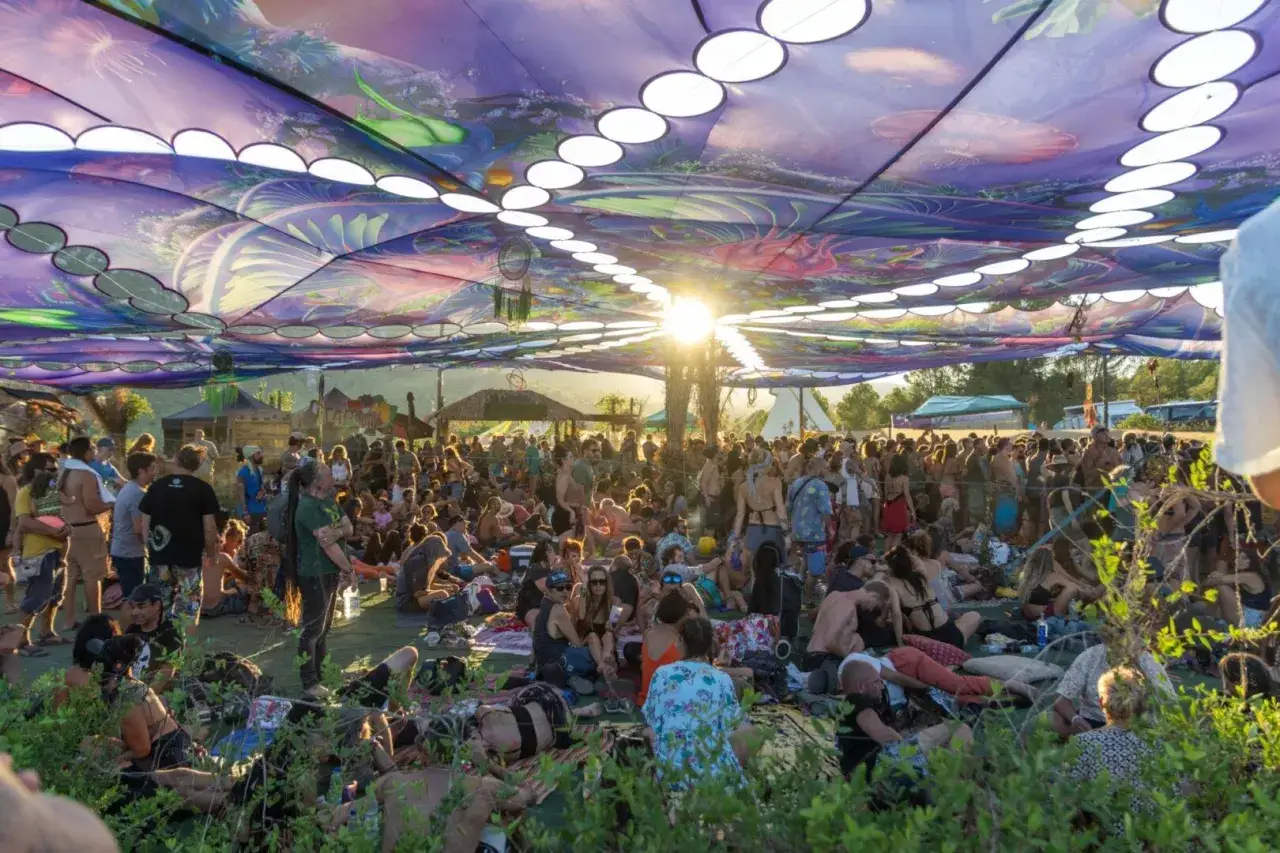 Multitud disfrutando bajo un toldo psicodélico en un festival psytrance España. El sol se filtra, creando un ambiente mágico.