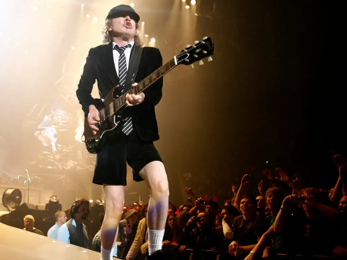 Gitarzysta AC/DC w krótkich spodenkach i czapce gra na scenie dla rozentuzjazmowanego tłumu.