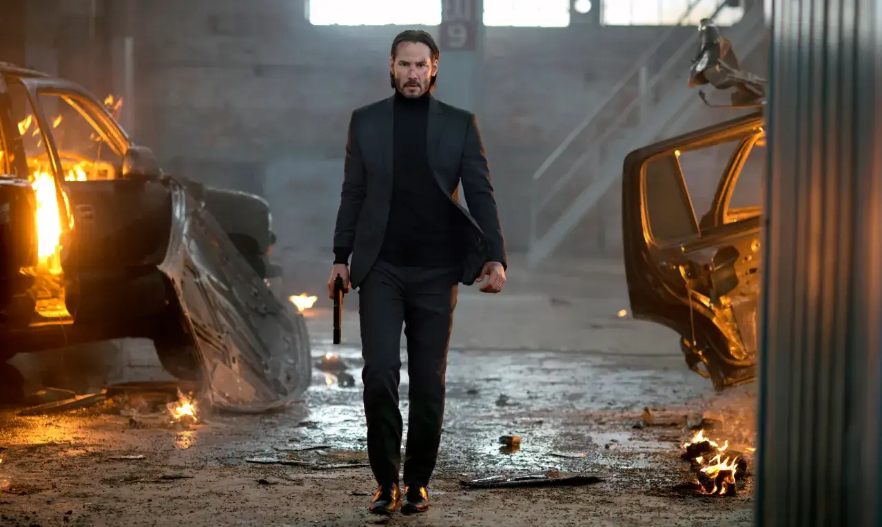 John Wick, med en pistol i handen, går genom en lagerlokal med brinnande bilvrak.