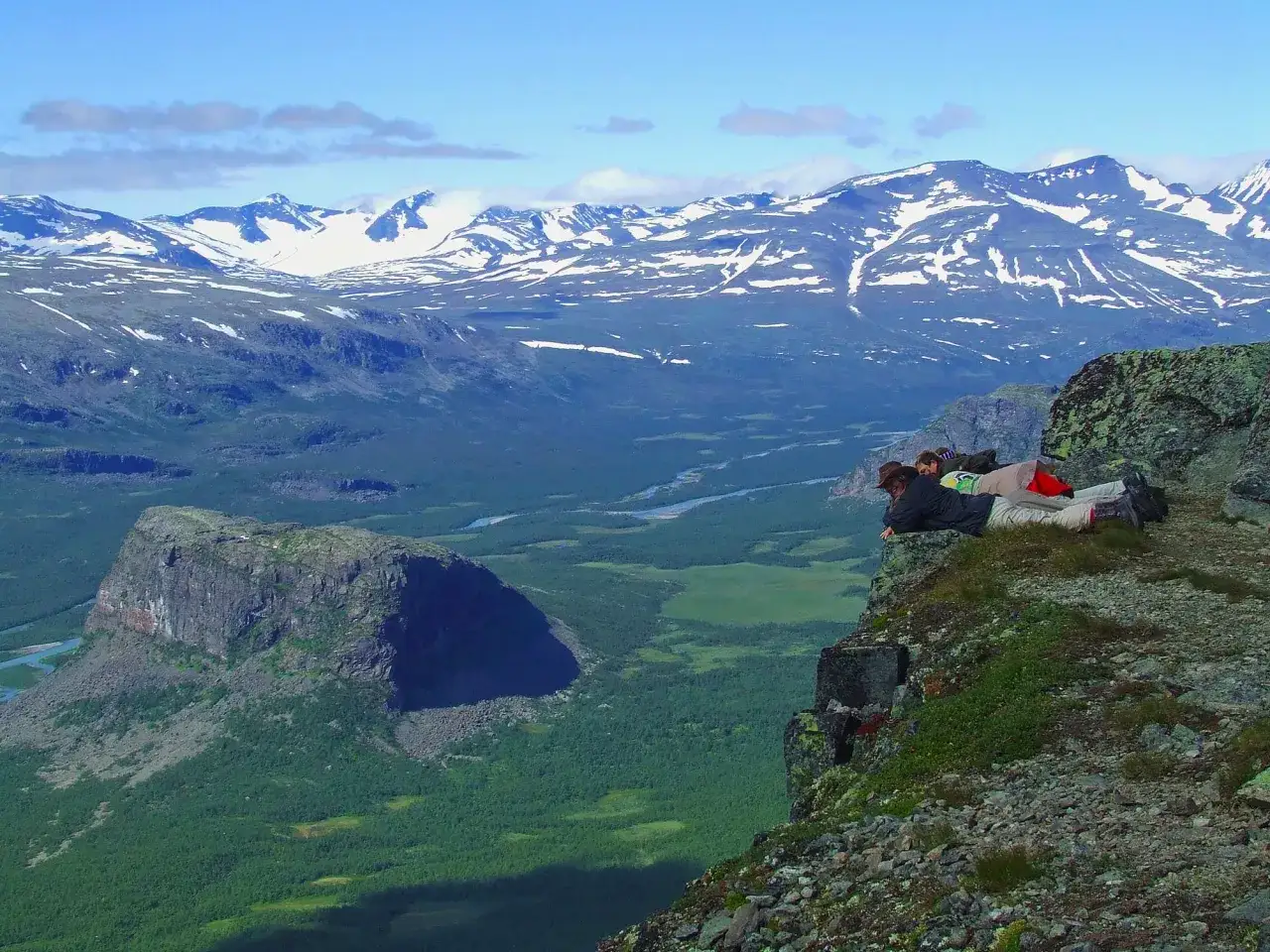 Zwei Wanderer ruhen sich auf einem Felsvorsprung aus und blicken auf ein weites Tal mit schneebedecktem Gebirge Schweden im Hintergrund.