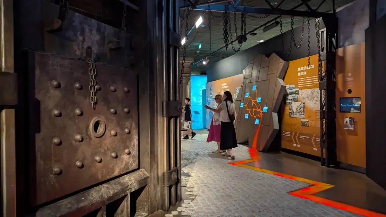 W muzeum śląskim zwiedzający oglądają wystawę o historii miasta. Widać masywne, metalowe drzwi i ekspozycję z mapami.