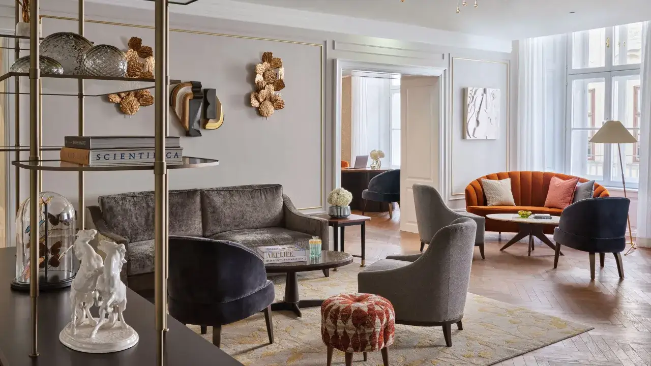 Salon élégant avec canapés velours, fauteuils gris, et un canapé orange vif. Des objets décoratifs dorés, dont des feuilles, ajoutent une touche de luxe. Le **bois de rose couleur** se retrouve dans les meubles et les détails.