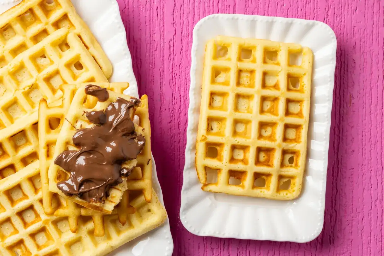 Una deliziosa ricetta waffle: waffle dorati su piatti bianchi, uno con crema al cioccolato, su uno sfondo rosa vibrante.