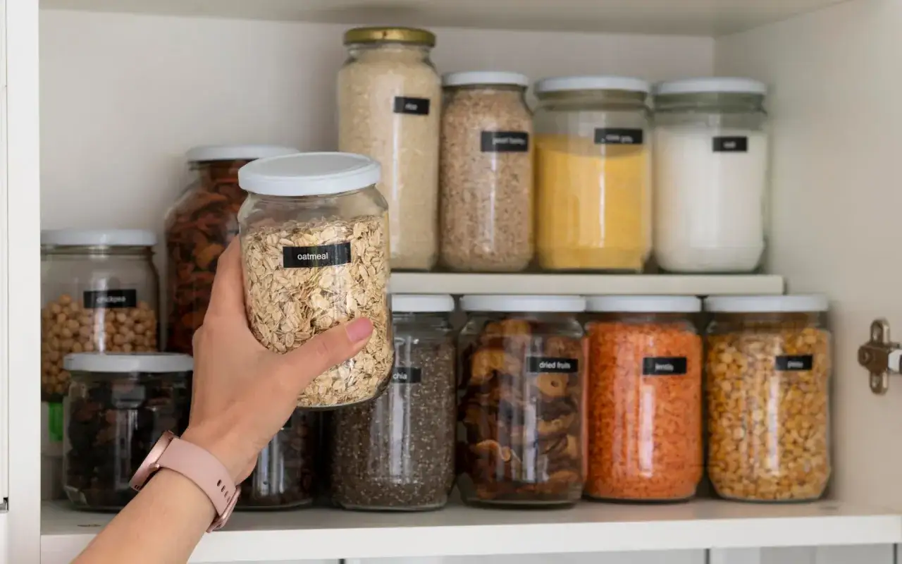 Mano toma un frasco de avena. Estante lleno de frascos con alimentos no perecederos: arroz, lentejas, garbanzos, chía, frutas secas.