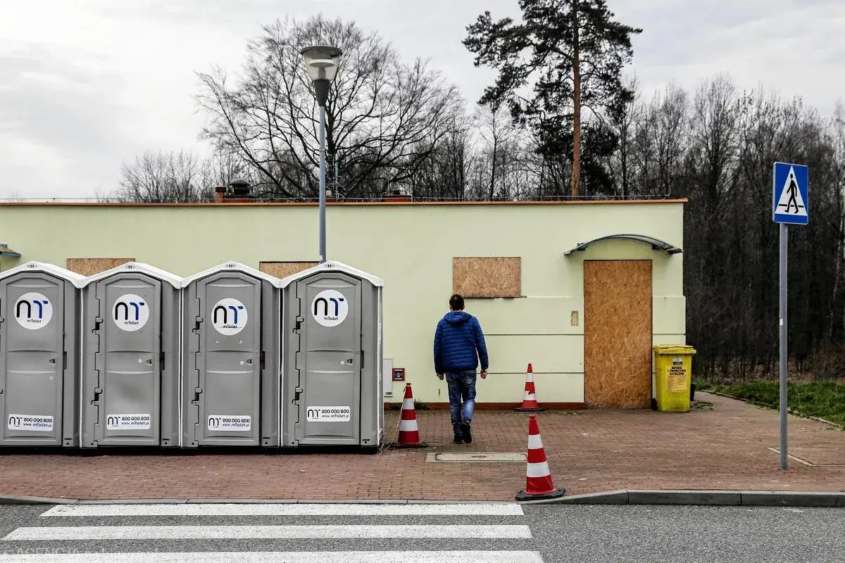 Cztery przenośne toalety mToilet stoją obok budynku. Mężczyzna w niebieskiej kurtce idzie w stronę budynku. Co to znaczy? Toalety na wynajem.
