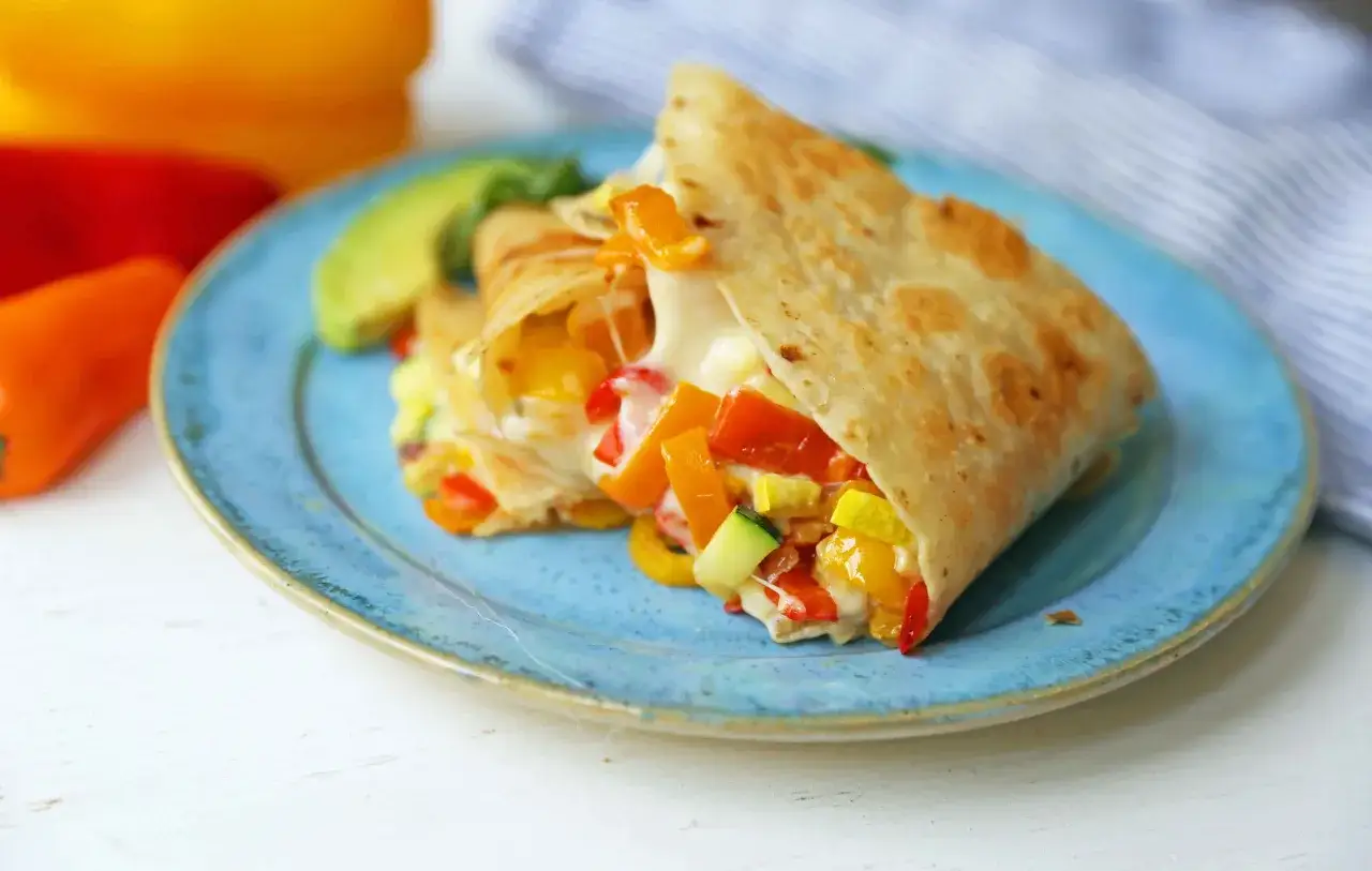 Leckeres Quesadillas Rezept mit geschmolzenem Käse, Paprika und Zucchini auf einem blauen Teller. Dazu Avocado und bunte Paprika.