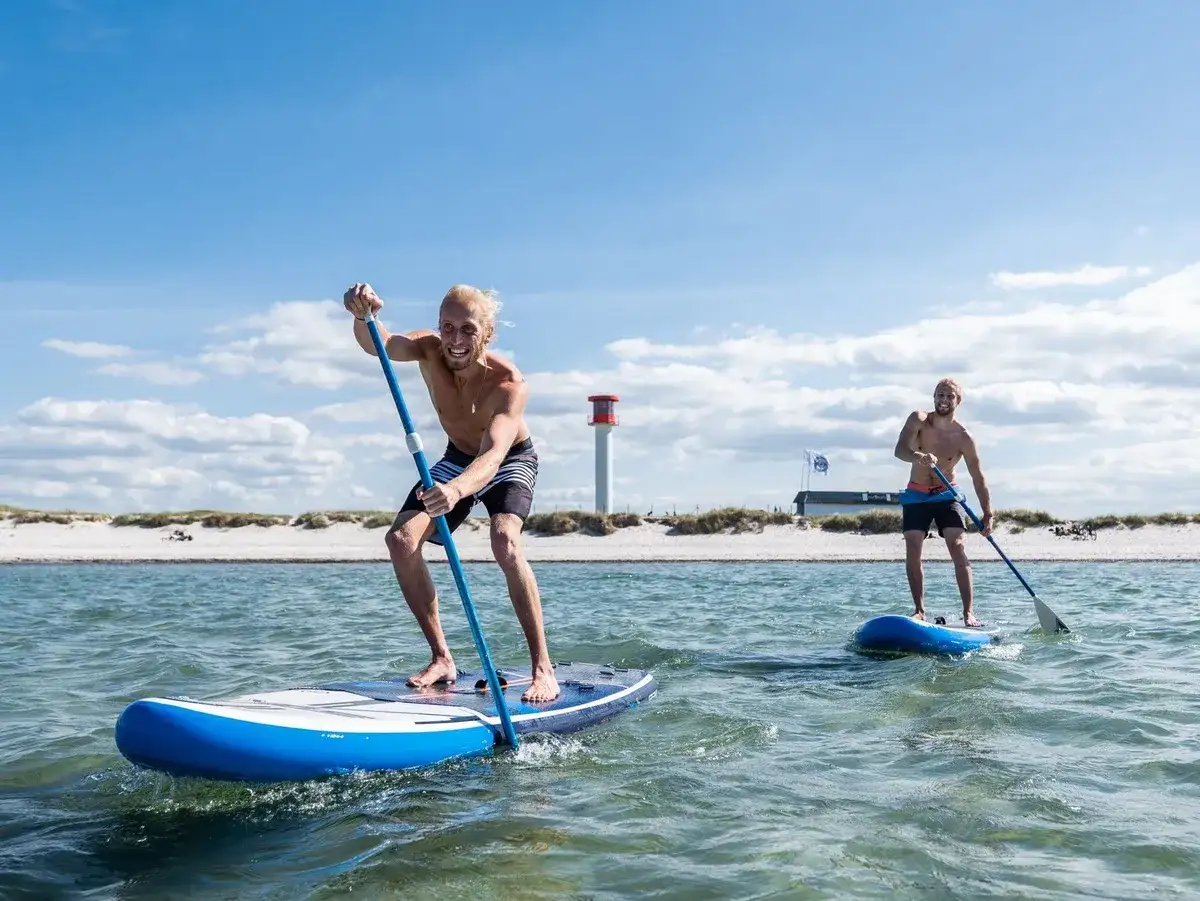 Zwei M&auml;nner genie&szlig;en SUP in Schleswig-Holstein. Sie paddeln auf dem Wasser, im Hintergrund ein Leuchtturm und Strand.