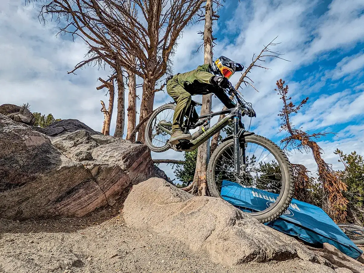 A mountain biker navigates a rocky trail on a mullet bike, with dry trees and a blue mat in the background under a cloudy sky.