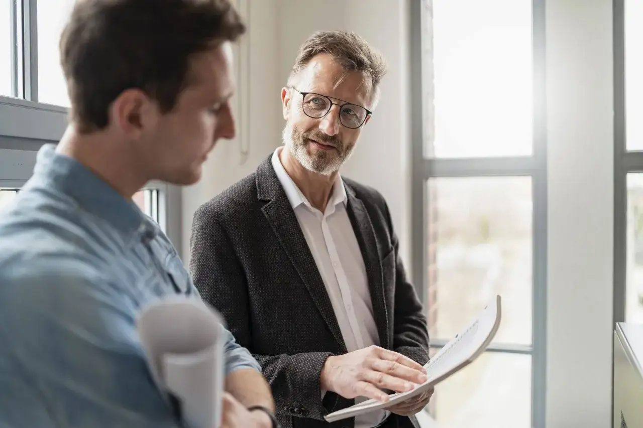 Zwei Männer besprechen Dokumente. Der ältere Mann mit Brille erklärt dem jüngeren, wie oft Gehalt verhandeln sinnvoll ist.