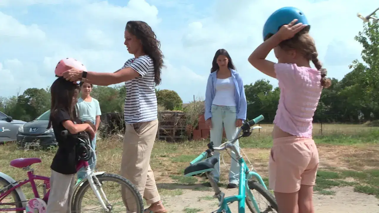 Une femme aide une jeune fille à mettre un casque rose pour faire du vélo. Une autre jeune fille met un casque bleu. Une belle journée pour devenir famille d'accueil pour les vacances.