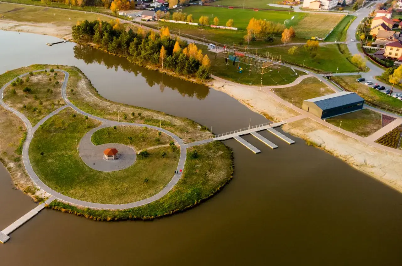 Malownicze, nisko jezioro z wyspą, altaną, ścieżkami i nowoczesnym budynkiem. W tle widać drzewa, plażę i zabudowania.