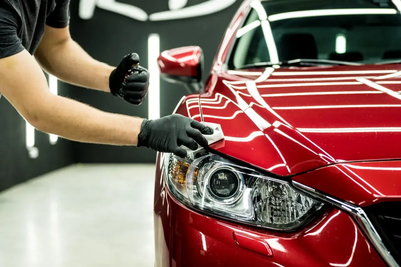 Hands in black gloves apply ceramic wax to a shiny red car headlight, protecting its finish.