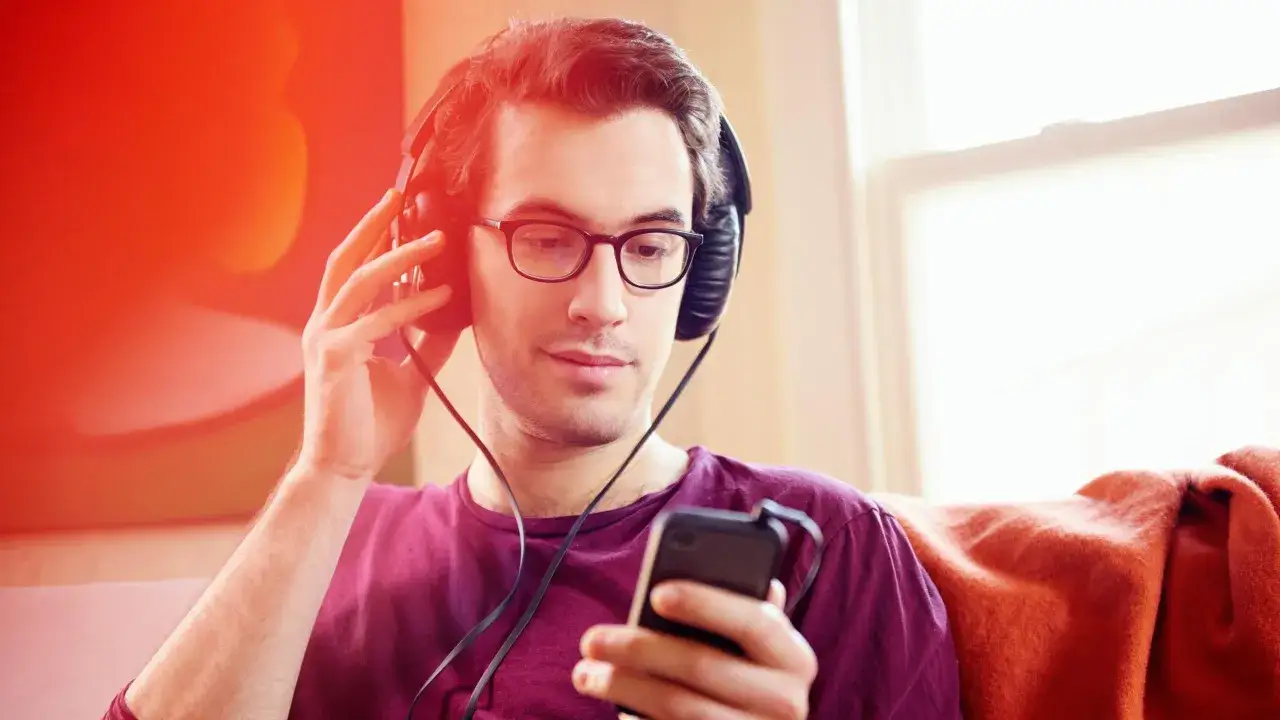 Hombre con auriculares y gafas escucha un podcast de psicolog&iacute;a en su tel&eacute;fono, aprendiendo y relaj&aacute;ndose en casa.
