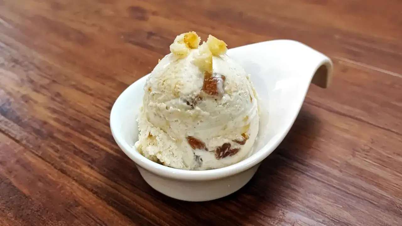 Ein Löffel cremiges Apfel-Eis mit Rosinen und Streuseln, serviert in einer weißen Schale auf Holztisch.