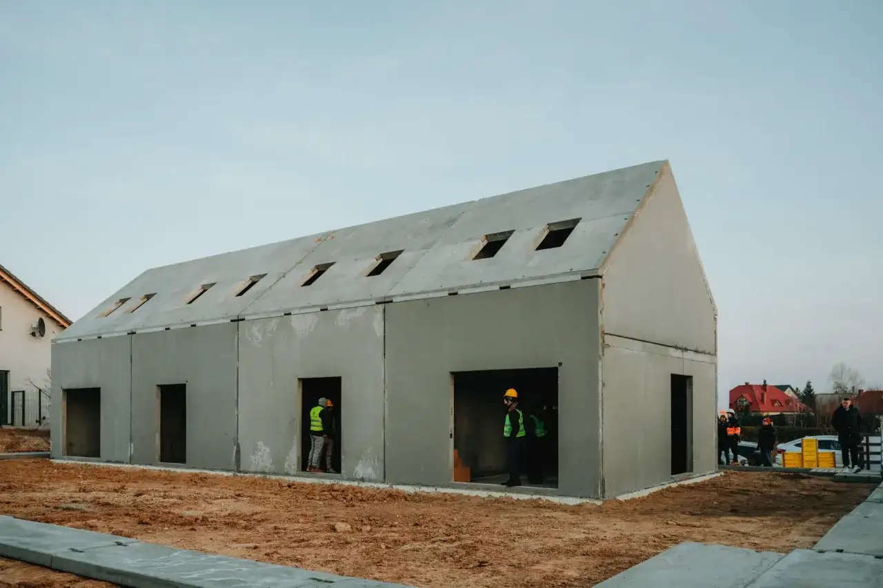 Budowa domu z prefabrykatów betonowych. Na placu budowy widać robotników w kamizelkach odblaskowych.