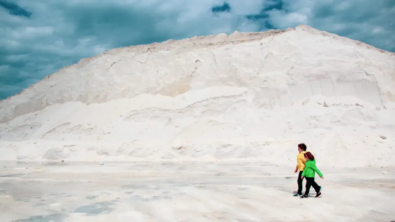 Due bambini passeggiano in un paesaggio lunare, un tavoliere delle Puglie dominato da una montagna di sale bianco sotto un cielo minaccioso.