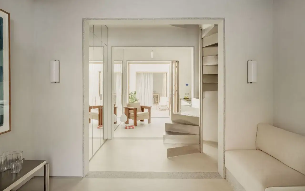 Un salón luminoso con un sofá beige, una escalera de caracol metálica y un sillón de madera. El espacio irradia un **timeless design** con sus líneas limpias y materiales naturales.