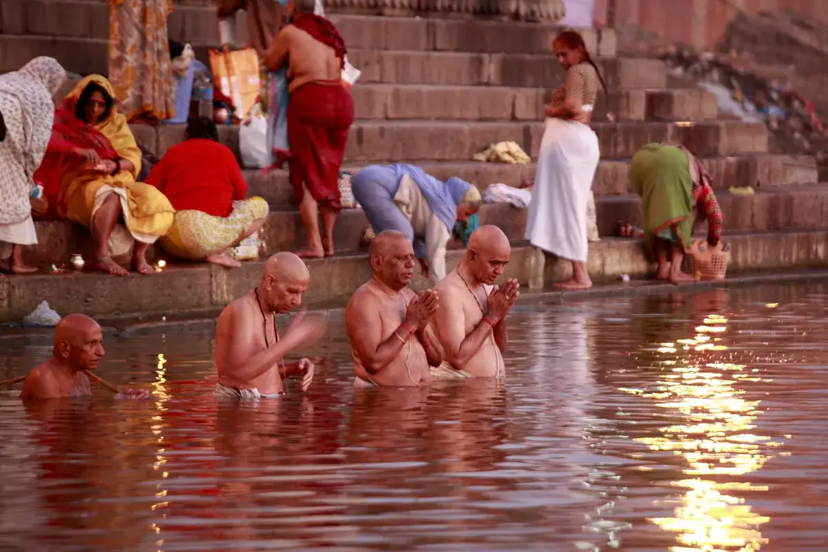 Hombres rezan en el r&iacute;o Ganges, con peregrinos en las escalinatas al fondo.