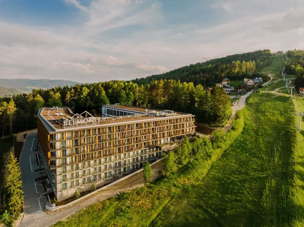 Nowoczesny hotel Belmonte Krynica w otoczeniu zielonych wzgórz i lasów, z widokiem na stok narciarski.