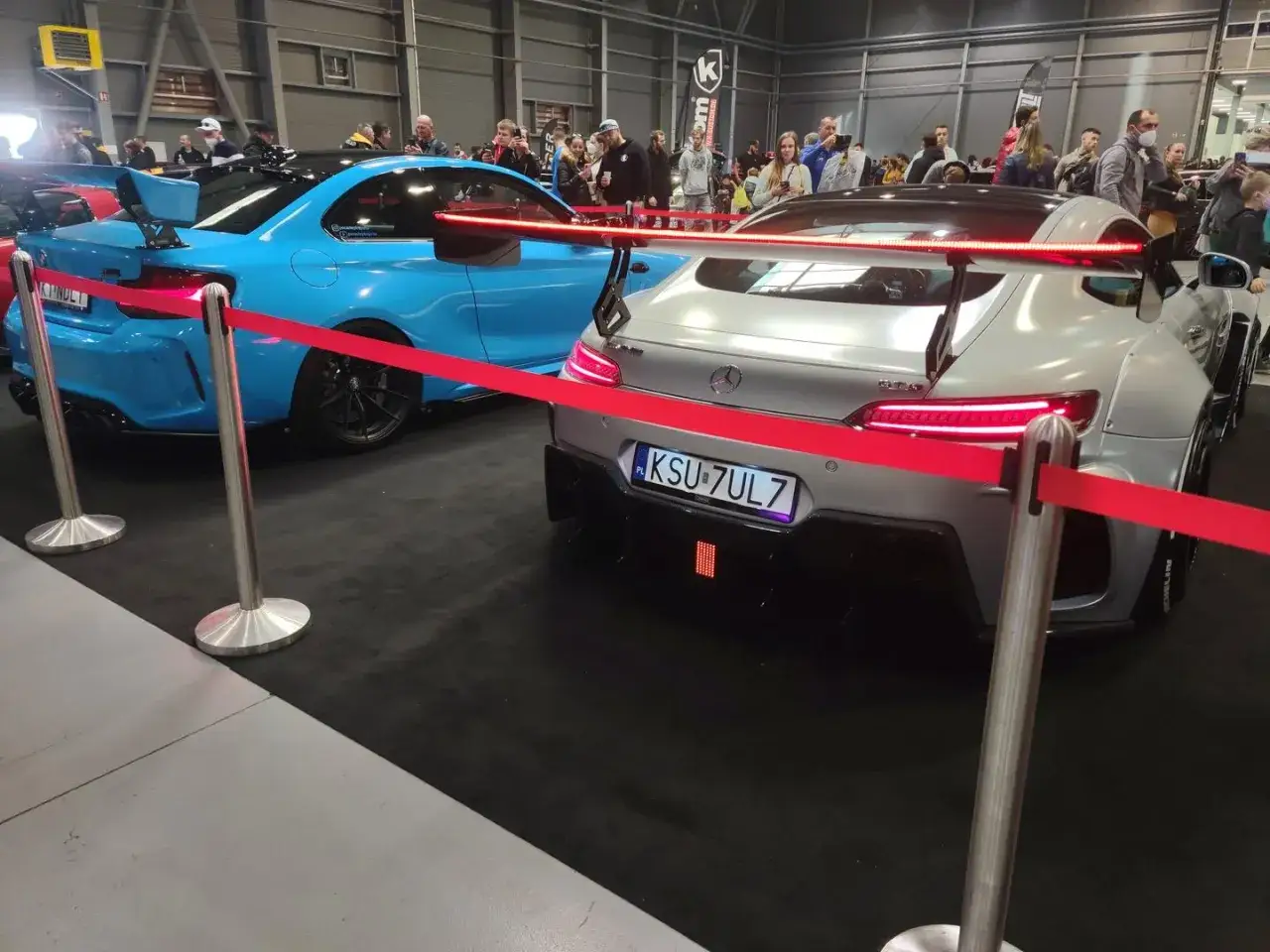 Na targach motoryzacyjnych, gdzie odbywa się ksu rejestracja, błyszczą dwa sportowe auta: niebieskie BMW i srebrny Mercedes-AMG GT.