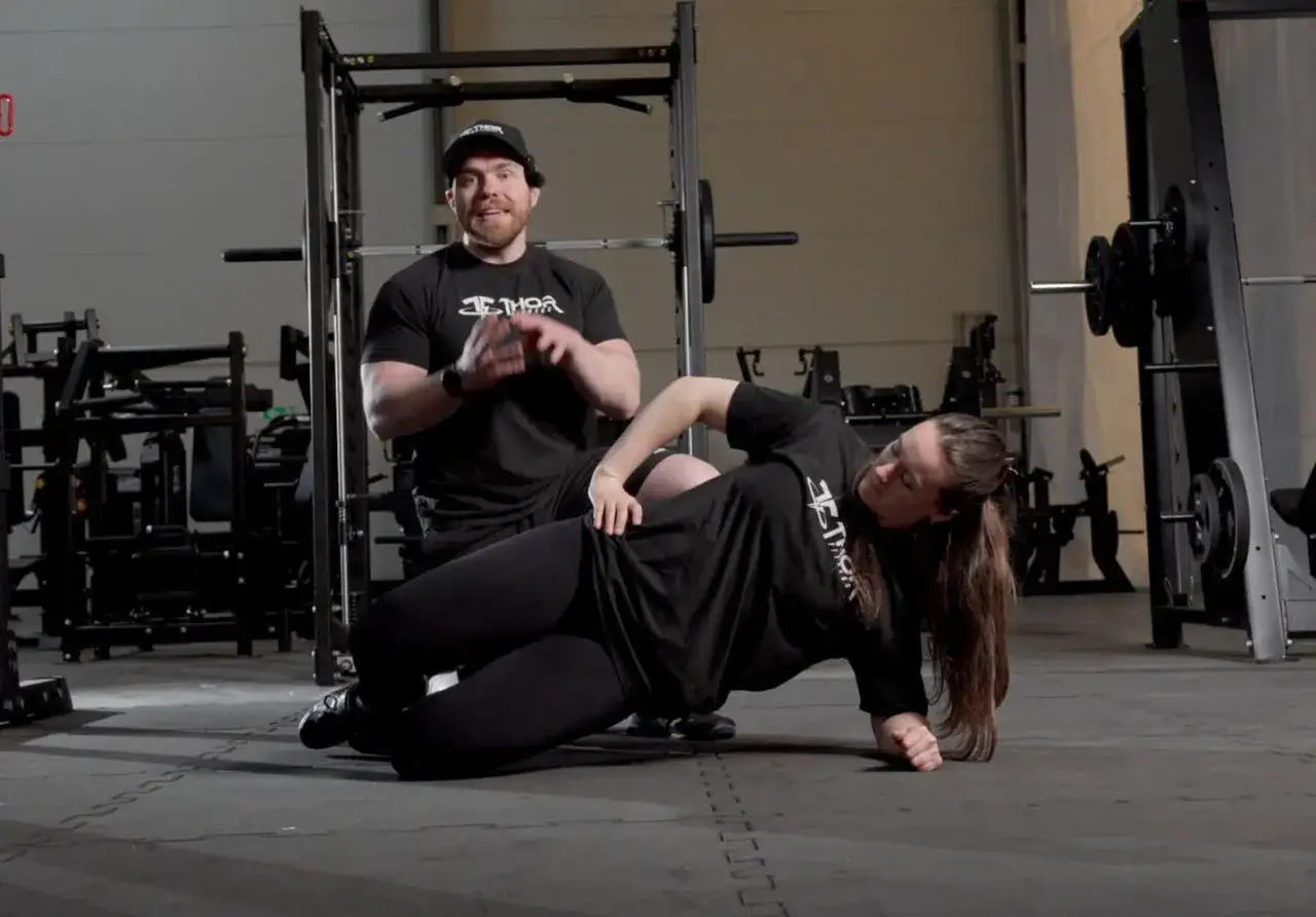 Man instruerar kvinna i sido planka p&aring; gymgolv.