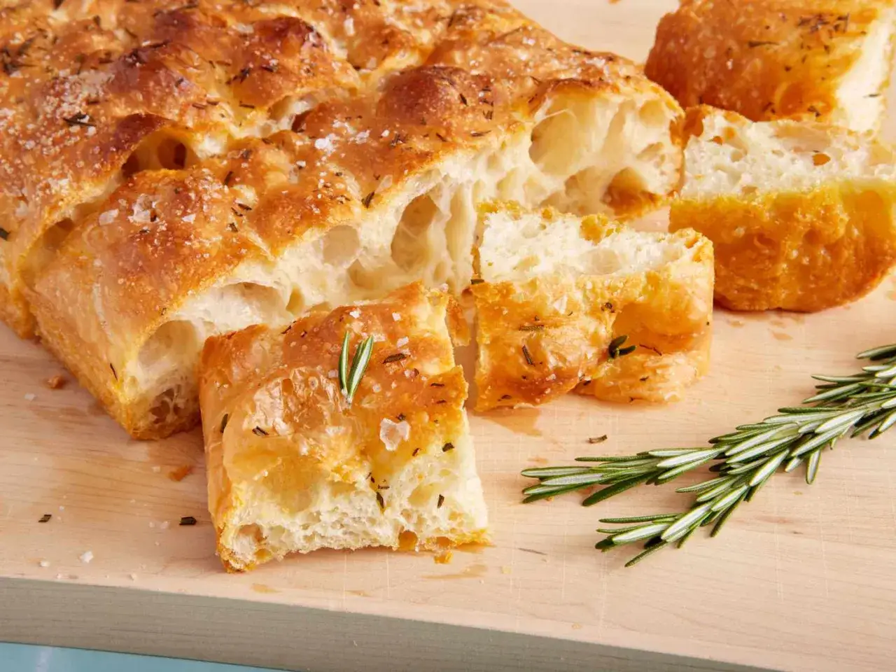 Focaccia pomodoro dorada y esponjosa, con romero fresco y sal gruesa, cortada en trozos sobre tabla de madera.