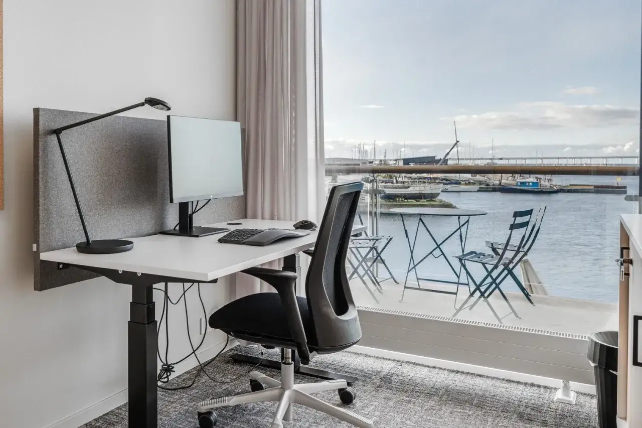 Ett modernt kontor med höj- och sänkbart skrivbord, dator och en fantastisk utsikt över en hamn. Perfekt för att förstå vad betyder hybrid jobb.