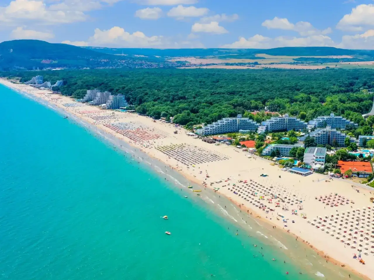 Słoneczne wakacje w Bułgarii: szeroka, piaszczysta plaża z mnóstwem leżaków i parasoli, turkusowe morze, hotele i zielone wzgórza w tle.