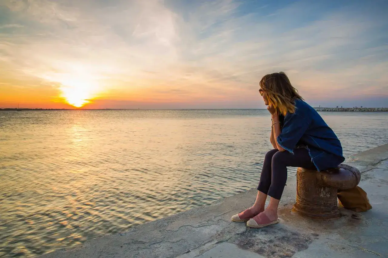 Donna seduta su un ormeggio osserva il tramonto a Rimini, con il sole che si riflette sul mare calmo.
