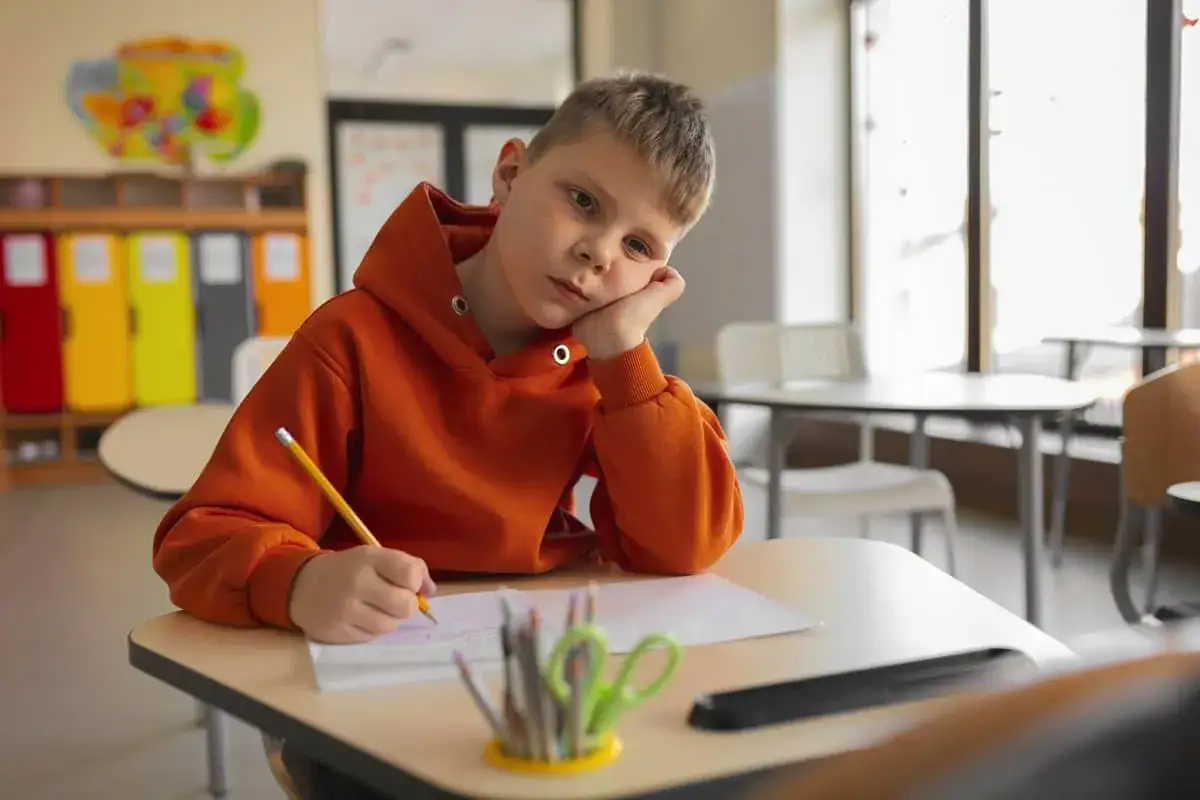Un niño con expresión triste en clase, apoyado en su mano, intentando escribir. Una señal de cómo detectar el bullying.
