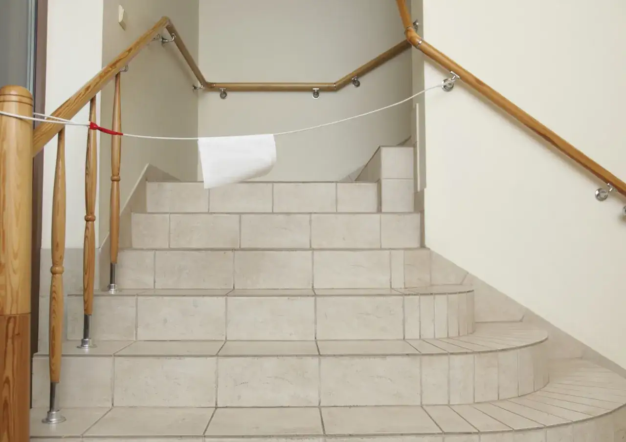 Escalier avec carrelage clair. Le carrelage de chaque marche ou contremarche est visible. Une corde blanche est tendue, avec un morceau de papier blanc.