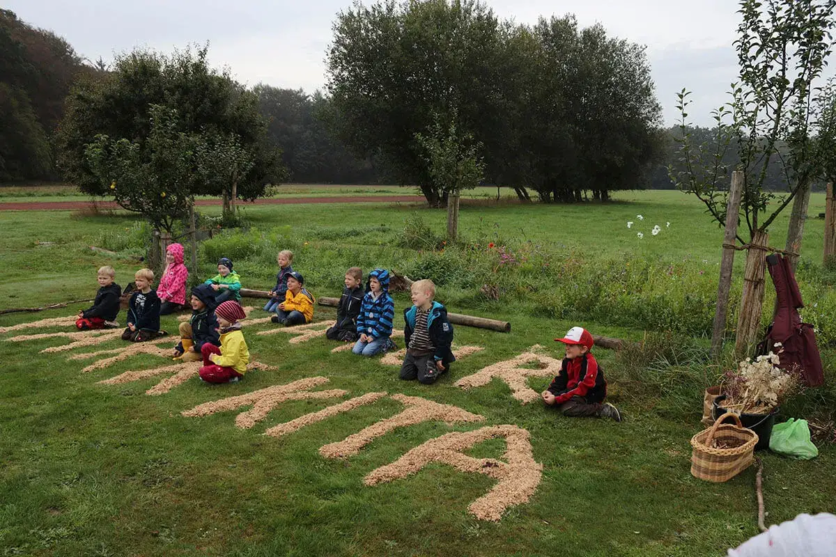 Kinder lernen im umweltschutz kindergarten spielerisch. Sie sitzen im Kreis auf einer Wiese, umgeben von B&auml;umen.