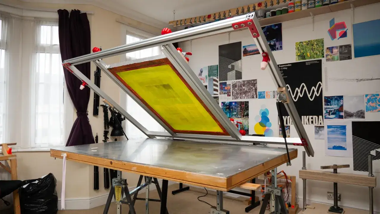 Mesa de trabajo con marco de serigraf&iacute;a y pantalla amarilla lista para imprimir. Paredes decoradas con obras de arte y materiales de serigraf&iacute;a.