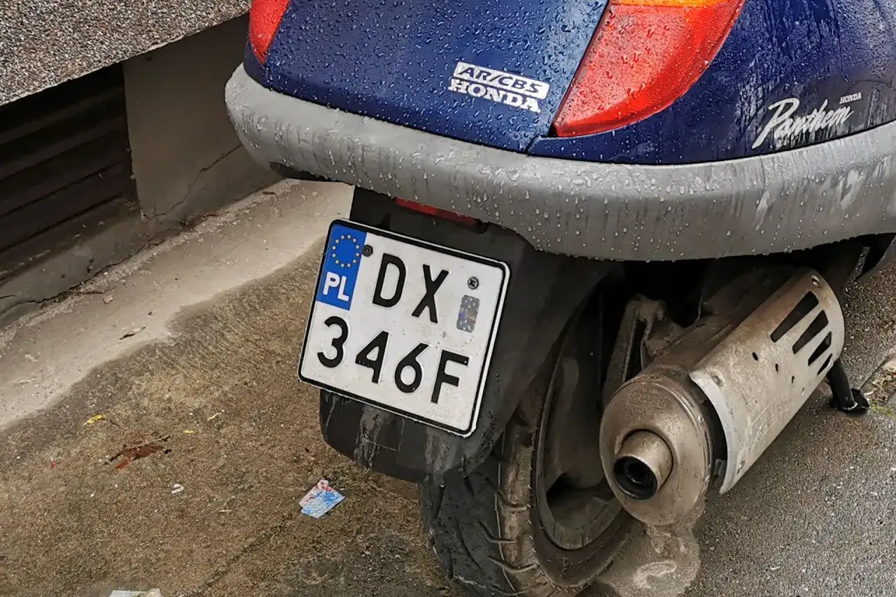 Niebieski skuter Honda Pantheon z tablicą rejestracyjną DX 346F stoi na mokrym chodniku.