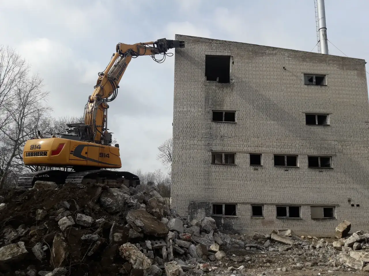 Żółta koparka Liebherr niszczy budynek z cegły. Wokół gruz. Sprawdź wyburzenia cena za m3.