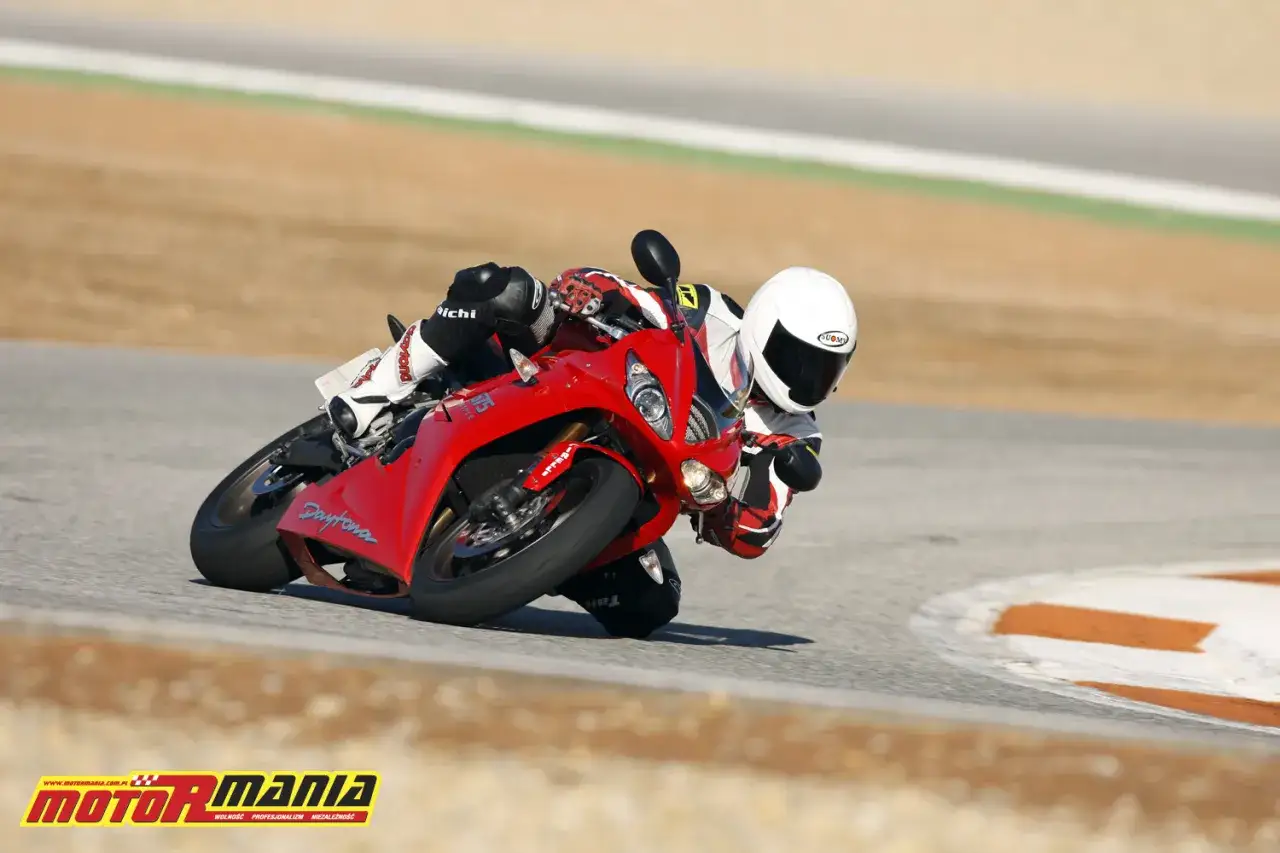 Motocyklista w kasku i kombinezonie pochyla się na torze, wykonując dynamiczną pozycję na motocyklu.