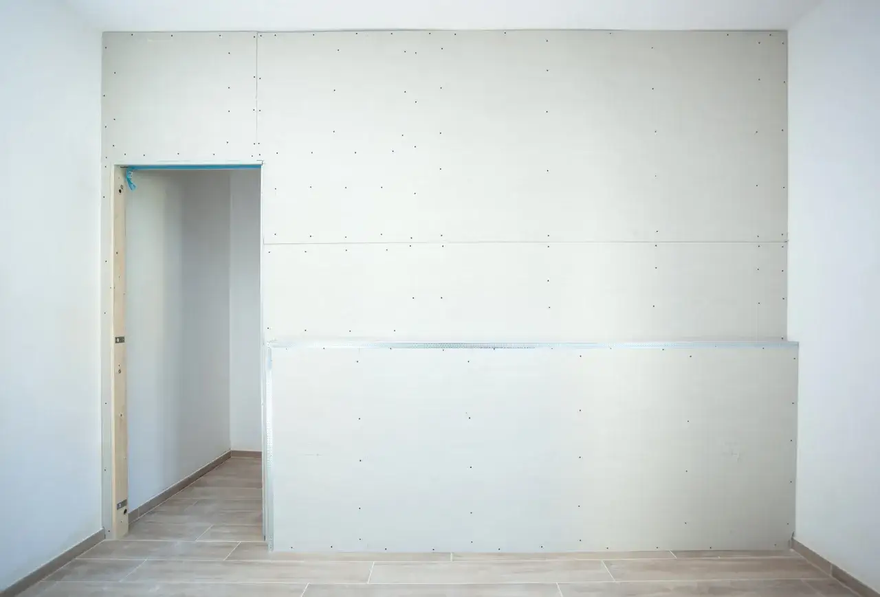 Un mur en plaques de plâtre prêt pour un enduit projeté intérieur. Une ouverture laisse entrevoir une pièce adjacente.