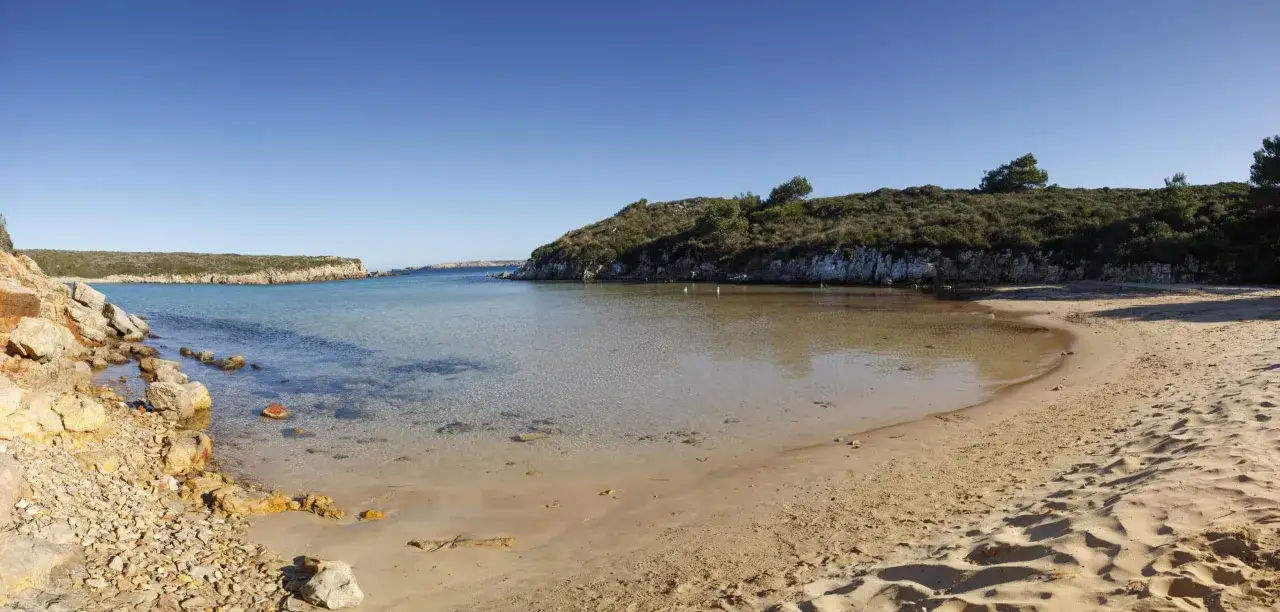 Una serena cala pudent con aguas cristalinas y arena dorada, rodeada de colinas cubiertas de vegetación y rocas.