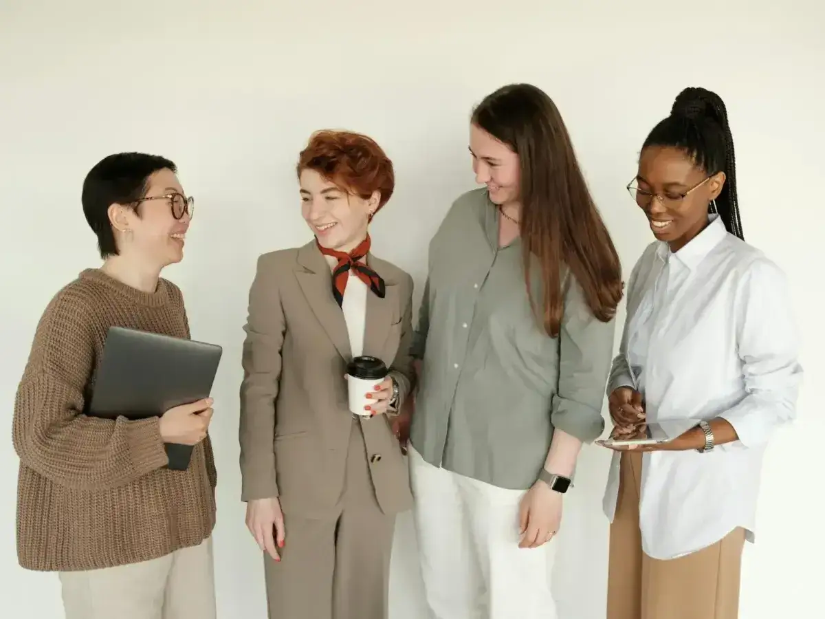 Quattro colleghe sorridono. Una con un tailleur sembra una collega che vuole comandare, tenendo una tazza.