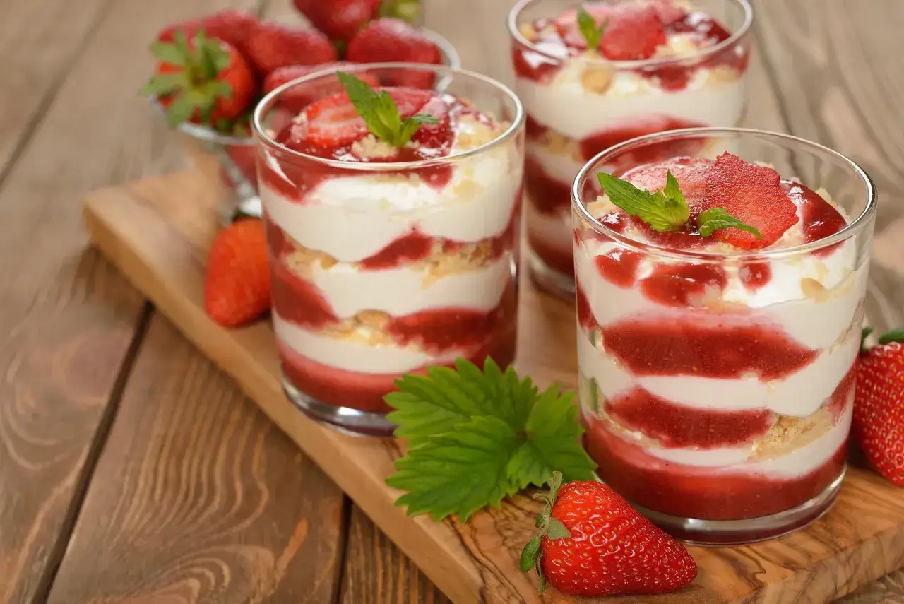 Trois verrines de tiramisu fraise, garnies de fruits rouges et de menthe, sur un plateau en bois.