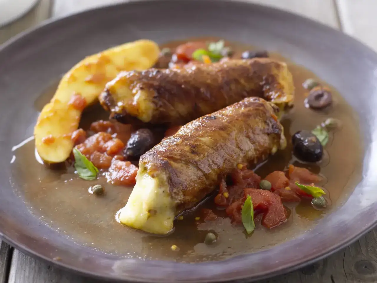 Zwei saftige Kalbsrouladen italienisch, gefüllt mit Käse, serviert mit Tomaten, Oliven und Kartoffeln in einer köstlichen Sauce.