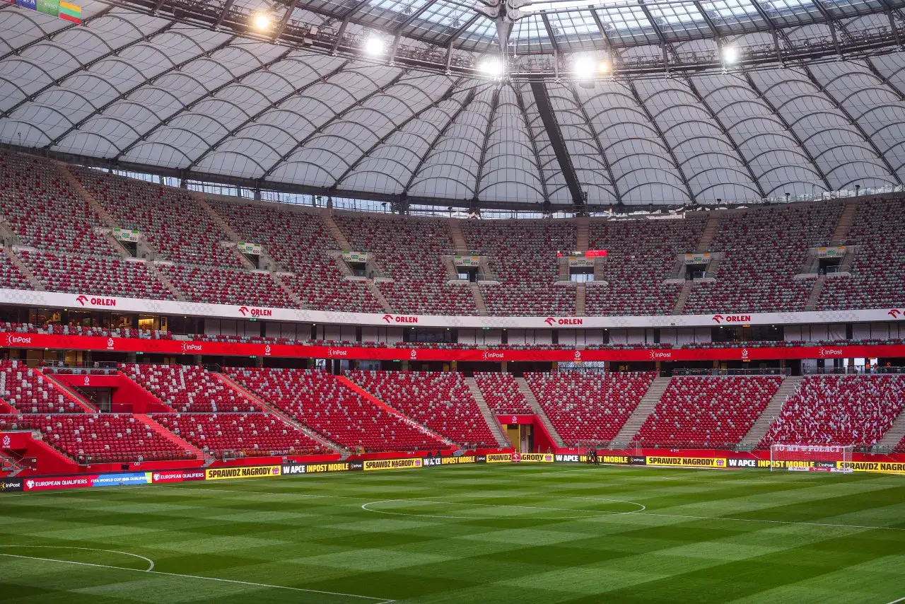 Pusta trybuna stadionu, gotowa na emocje pierwszej ligi piłki nożnej. Czerwono-szare krzesełka czekają na kibiców.
