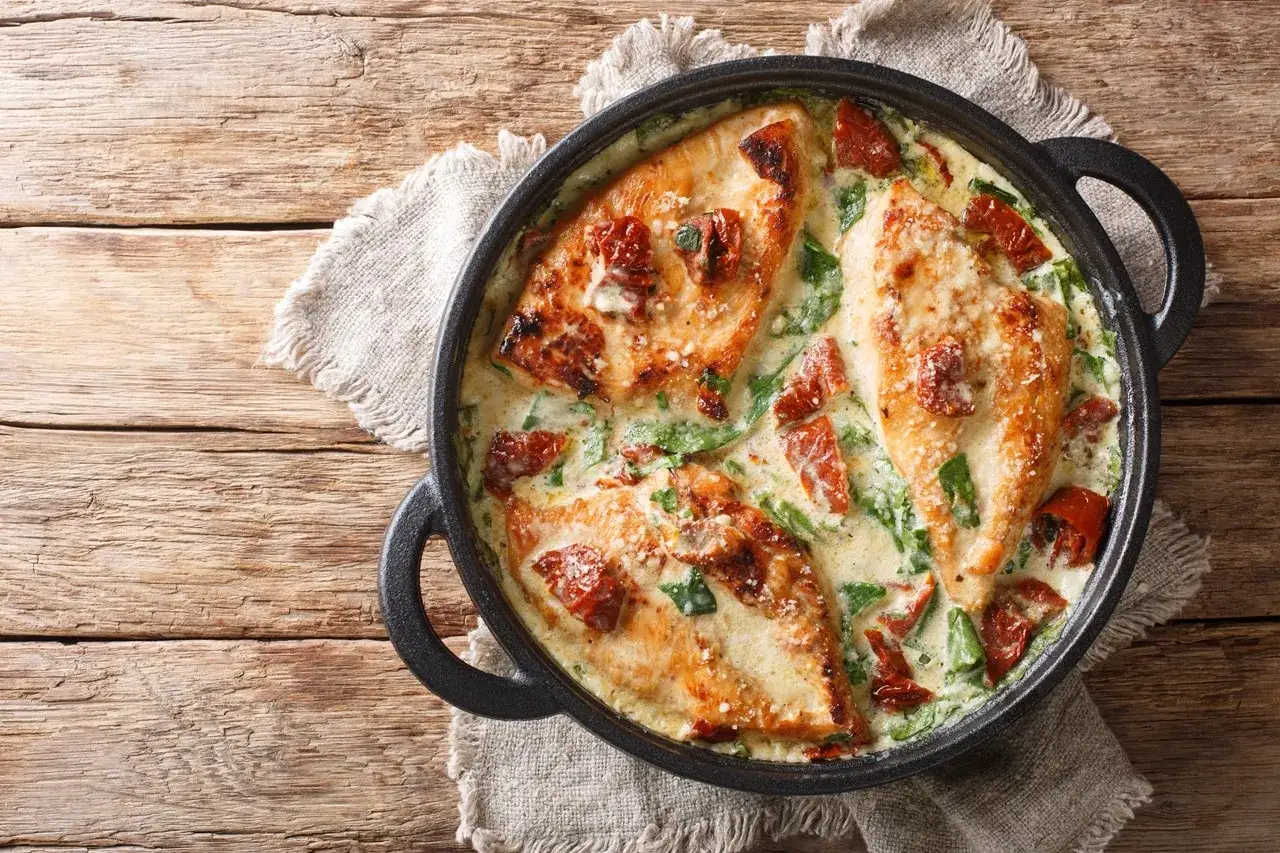 Trois morceaux de blanc de poulet dorés baignent dans une sauce crémeuse aux épinards et tomates séchées, servis dans une poêle en fonte sur une table en bois.