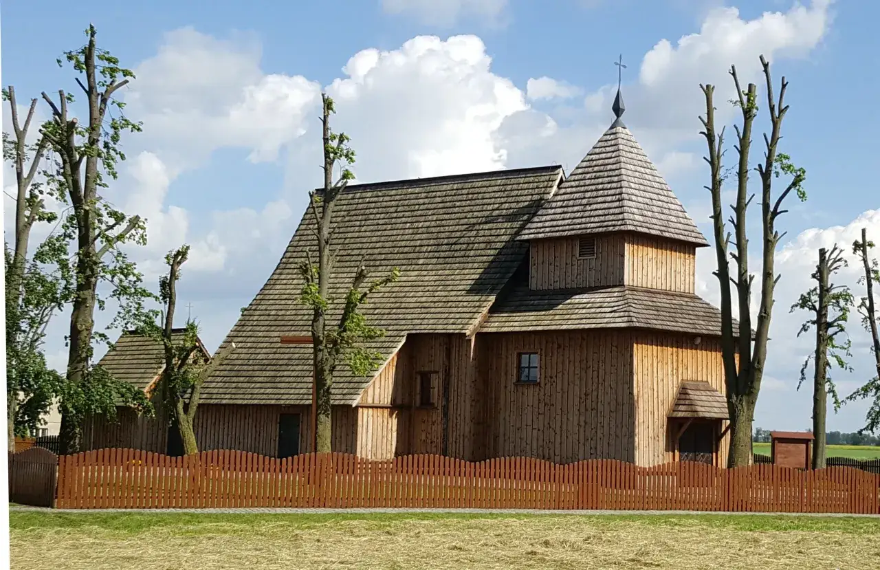 Drewniany kościółek w Nowej Wsi Królewskiej, otoczony drewnianym płotem i przyciętymi drzewami, pod błękitnym niebem z chmurami.