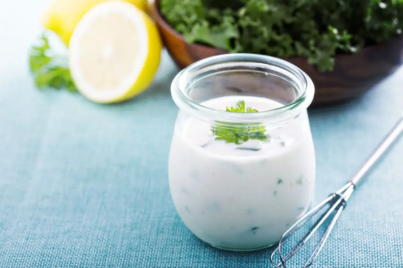 Cremige Joghurt-Mayonnaise mit Kräutern in einem Glas, daneben Zitronen und Grünkohl.