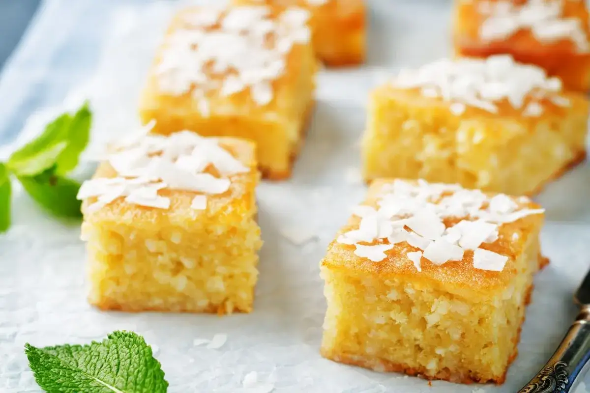 Fette quadrate di torta al cocco, guarnite con scaglie di cocco e foglie di menta fresca.