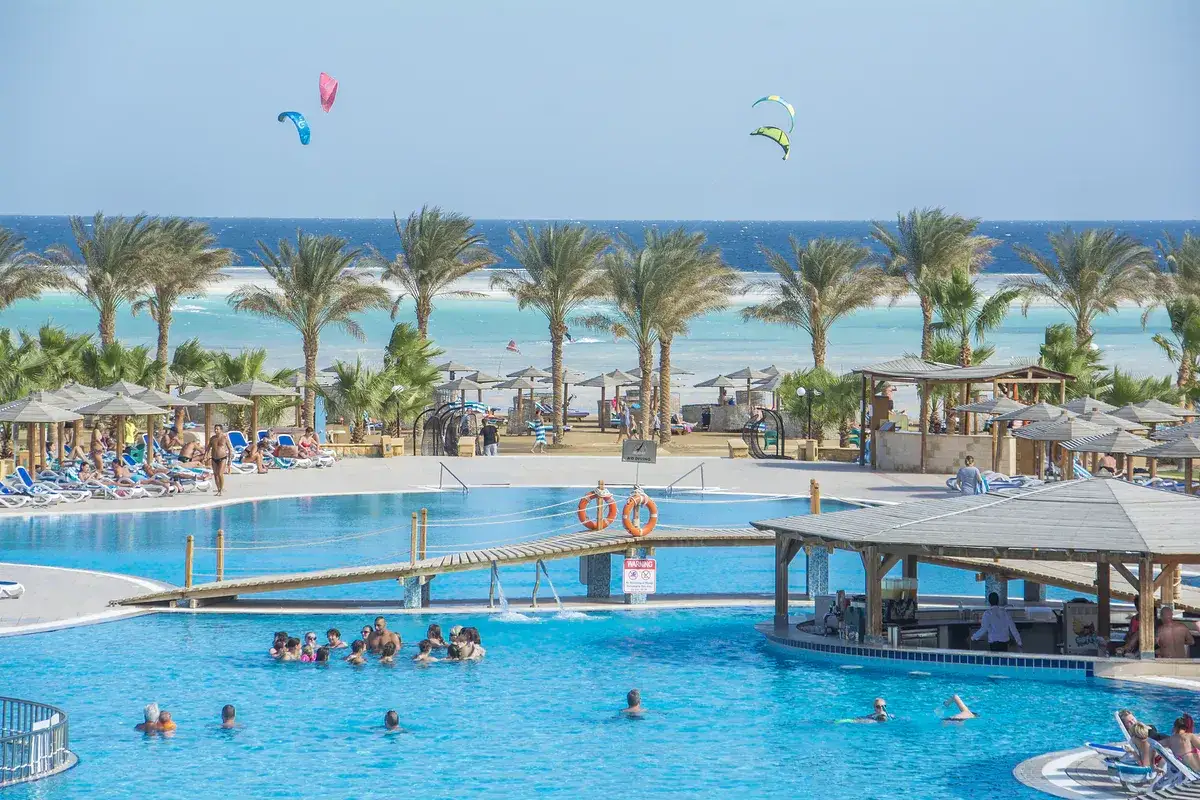 Słoneczna pogoda w Egipcie, Marsa Alam. Ludzie kąpią się w basenie, odpoczywają na leżakach pod palmami, a na niebie latają kolorowe latawce.